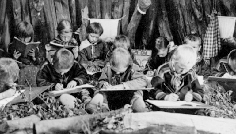 interiör från skolkåta med barn som sitter på björkris och skriver på skrivtavlor